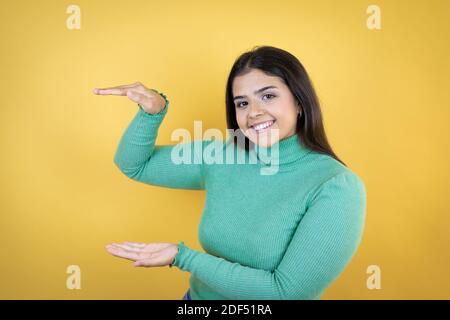 Junge kaukasische Frau über isolierten gelben Hintergrund gestikulieren mit Händen zeigt große und große Größe Zeichen, Maßnahme Symbol. Lächelnd. Messkonep Stockfoto