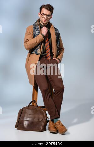 Volle Länge der stilvollen Mann im Winter Mantel und Brille Anpassen der Krawatte, während Sie auf dem Stuhl mit der Hand in der Tasche sitzen In der Nähe Aktentasche auf grau Stockfoto