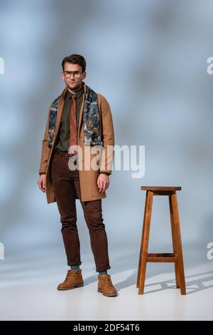 Volle Länge der stilvollen Mann im Winter Outfit und Brille Steht in der Nähe von Holzhocker auf grau Stockfoto