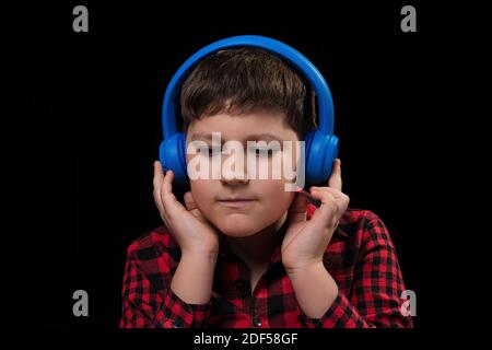 Niedlichen hübschen Jungen Teenager in rot kariert Hemd aufmerksam begeistert Hören Sie Musik in großen blauen kabellosen Ohrhörern Isoliert schwarz b Stockfoto