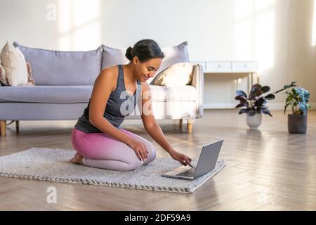 Lächelnde afroamerikanische Frau, die sich zu Hause vor einem Laptop-Monitor auf ein Workout vorbereitet. Sie sitzt auf dem Boden vor dem Laptop. Stockfoto