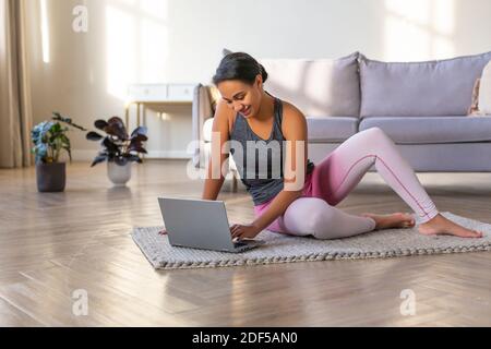 Lächelnde afroamerikanische Frau, die sich zu Hause vor einem Laptop-Monitor auf ein Workout vorbereitet. Sie sitzt auf dem Boden mit Laptop. Stockfoto