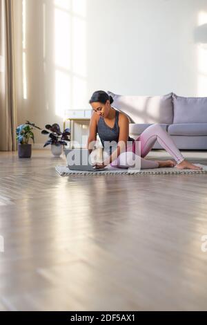 Lächelnde afroamerikanische Frau, die sich zu Hause vor einem Laptop-Monitor auf ein Workout vorbereitet. Sie sitzt auf dem Boden mit Laptop. Vertikales Foto. Stockfoto