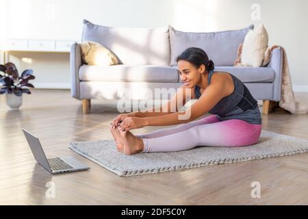 Lächelnde afroamerikanische Frau macht Stretching zu Hause. Sie sitzt auf einer Trainingsmatte vor einem Laptop-Monitor. Stockfoto