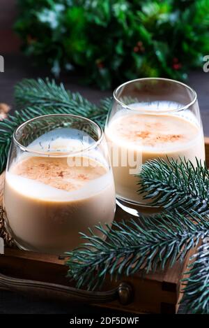 Eierwein Weihnachtsmilch-Cocktail mit Zimt, serviert in zwei Gläsern auf Vintage-Tablett mit Tannenzweig auf dunklem Holzhintergrund Stockfoto