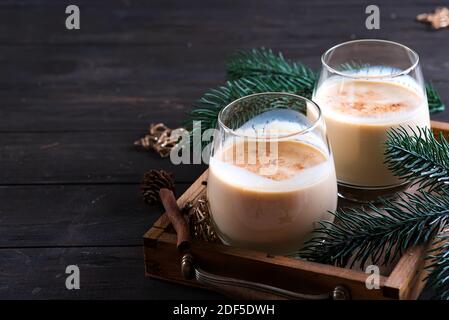 Eierwein Weihnachtsmilch-Cocktail mit Zimt, serviert in zwei Gläsern auf Vintage-Tablett mit Tannenzweig auf dunklem Holzhintergrund Stockfoto
