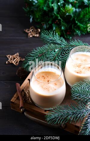 Eierwein Weihnachtsmilch-Cocktail mit Zimt, serviert in zwei Gläsern auf Vintage-Tablett mit Tannenzweig auf dunklem Holzhintergrund Stockfoto