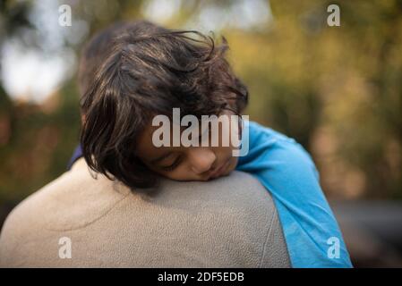 Ein indischer Brünette Vater mit seinem kleinen Jungen in seinem Schoß im Winternachmittag sitzt auf Holzstämmen im Wald Hintergrund. Stockfoto