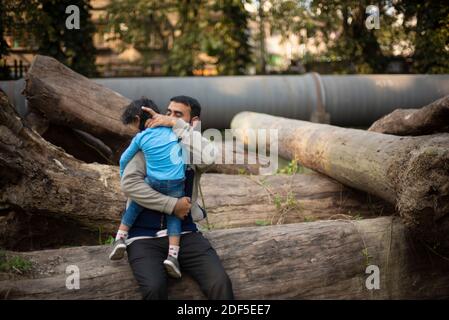 Ein indischer Brünette Vater mit seinem kleinen Jungen in seinem Schoß im Winternachmittag sitzt auf Holzstämmen im Wald Hintergrund. Stockfoto