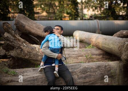 Ein indischer Brünette Vater mit seinem kleinen Jungen in seinem Schoß im Winternachmittag sitzt auf Holzstämmen im Wald Hintergrund. Stockfoto