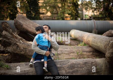 Ein indischer Brünette Vater mit seinem kleinen Jungen in seinem Schoß im Winternachmittag sitzt auf Holzstämmen im Wald Hintergrund. Stockfoto