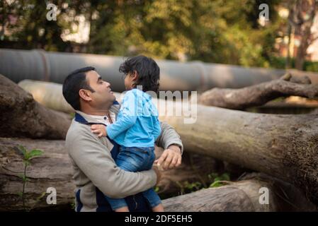 Ein indischer Brünette Vater mit seinem kleinen Jungen in seinem Schoß im Winternachmittag sitzt auf Holzstämmen im Wald Hintergrund. Stockfoto