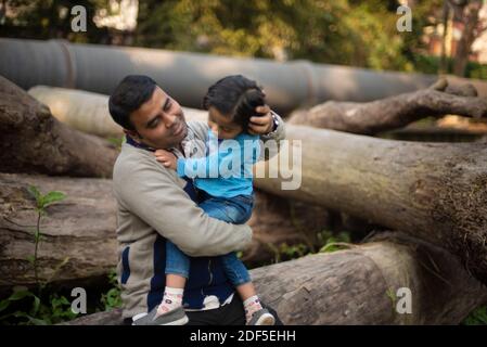 Ein indischer Brünette Vater mit seinem kleinen Jungen in seinem Schoß im Winternachmittag sitzt auf Holzstämmen im Wald Hintergrund. Stockfoto