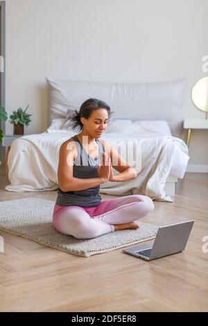 Vertikales Foto einer afroamerikanischen Frau meditiert zu Hause vor einem Laptop-Monitor mit Online-Tutorials. Stockfoto