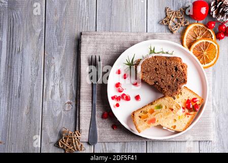Weihnachten in Scheiben geschnittene Schokoladenkuchen und Obstkuchen mit weißer Vereisung und Granatapfelkernen auf Teller mit Dekoration auf einem hölzernen grauen Hintergrund, flach legen Stockfoto