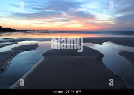 Eine lange Belichtung fängt Bewegung in den Wolken ein, während die aufgehende Sonne den Himmel mit Farbe füllt. Gezeitenbecken im dunklen Sand reflektieren das Licht der Dämmerung. Stockfoto