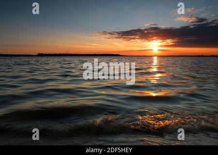 Die Sonne füllt den Horizont mit warmem, goldenem Licht, wenn sie durch dicke Wolken untergeht. Das Sonnenlicht funkelt auf den sanften, plätschernden Wellen eines ruhigen Meeres. Stockfoto