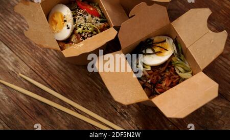 Chinesische Nudeln mit Huhn und Gemüse in Pappkartons auf einem hölzernen Hintergrund, asiatische Lebensmittelzustellung, Konzept von Street Food, Kopierraum Stockfoto