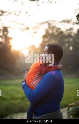 Indischer Telugu Brünette Vater und sein Baby im Winter Kleidungsstücke, die sich am Winternachmittag auf grünem Gras erfreuen Feld im Waldhintergrund Stockfoto