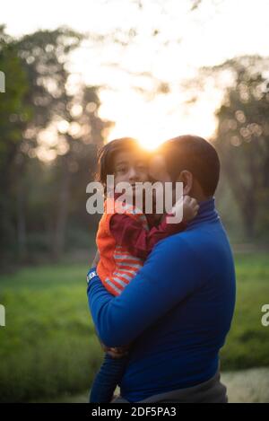 Indischer Telugu Brünette Vater und sein Baby im Winter Kleidungsstücke, die sich am Winternachmittag auf grünem Gras erfreuen Feld im Waldhintergrund Stockfoto