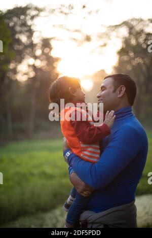 Indischer Telugu Brünette Vater und sein Baby im Winter Kleidungsstücke, die sich am Winternachmittag auf grünem Gras erfreuen Feld im Waldhintergrund Stockfoto