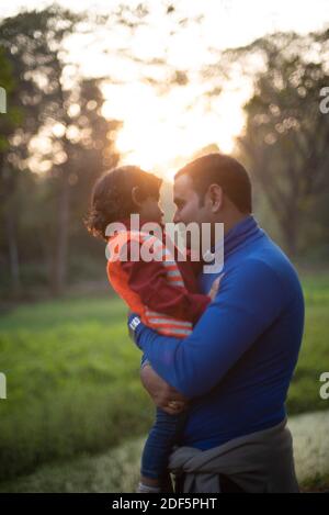 Indischer Telugu Brünette Vater und sein Baby im Winter Kleidungsstücke, die sich am Winternachmittag auf grünem Gras erfreuen Feld im Waldhintergrund Stockfoto