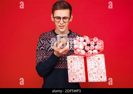 Ein Mann in einer Brille, mit einem Geschenk in der Hand und einem Blumenstrauß, schaut auf den Telefonbildschirm, auf einem roten Hintergrund. Frauentag Stockfoto