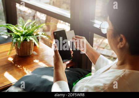 Ältere Dame sitzt auf einem Stuhl und hält die Kreditkarte mit Smartphone, Online-Shopping-Konzept Stockfoto