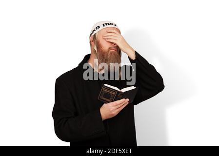 Frommer jüdischer Mann, der Siddur in seiner Hand hält, die Augen mit der Hand bedeckt, die auf weißem Hintergrund isoliert ist. Bärtiger Jude chassid mit Seitenschwänzen in weißem Kippach Stockfoto