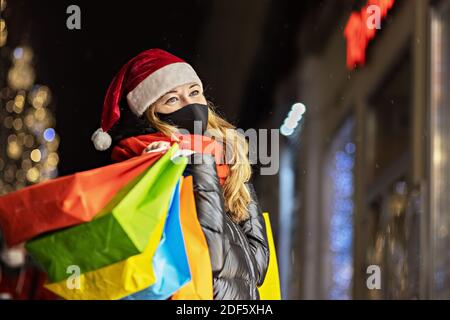 Eine Frau in einer schwarzen medizinischen Schutzmaske in einem Weihnachtsmann-Hut am Fenster eines Stadtladens mit Einkäufen in bunten Papiertüten. Neujahrsbummel Stockfoto