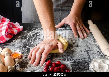 Hände kochen Teig kneten, ein Stück Teig mit Weizenmehl bestreuen. Low-Key-Schuss, Nahaufnahme der Hände, einige Zutaten rund um den Tisch Stockfoto