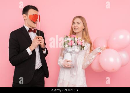 Ein liebevolles Paar, ein Mann hält zwei Papierherzen in den Augen, und eine Frau hält einen Blumenstrauß und Luftballons im Studio auf einem rosa Hintergrund Stockfoto