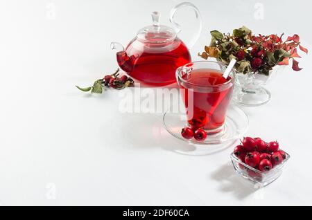 Herbstlicher roter Kräutertee in transparenter Teekanne, Tasse mit trockenen Blättern, Hagebutte, Weißdornbeeren im Sonnenlicht auf weißem Holztisch, Kopierraum. Stockfoto