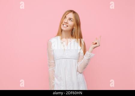 Ein glückliches, nachdenkliches Mädchen in einem weißen Kleid denkt über etwas nach, macht Pläne und zeigt auf einen leeren Raum im Hintergrund, auf einem rosa Backgr Stockfoto