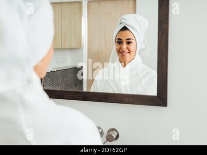 Frau im Bademantel, die ihre Reflexion anschaut. Schöne Brünette mit einem weißen Handtuch auf dem Kopf. Stockfoto