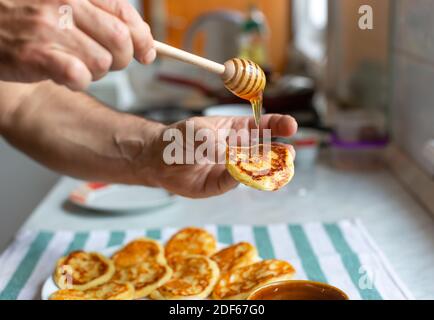 Mann gießt Honig auf rodigen hausgemachten Pfannkuchen. Honig tropft aus dem Honigstock auf leckeren Fritter Stockfoto