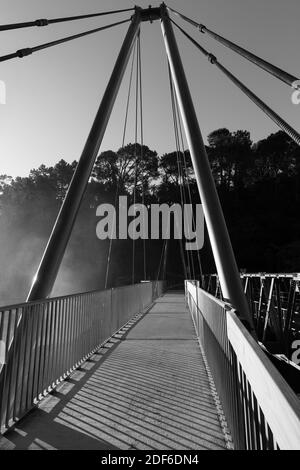 Eine Fußgängerhängebrücke, die die letzten Sonnenstrahlen einfängt. Spray aus einem Wasserfall driftet von links in das Foto. Schwarz und Weiß Stockfoto