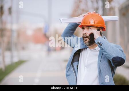 Junger Architekt hält Handy und Blaupause, während er wegschaut. Stockfoto