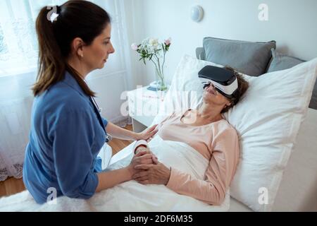 Ältere Patientin, die sich im Krankenhausbett entspannt, mit Virtual Reality-Headset. Startseite Krankenschwester sitzen auf dem Bett Blick auf ältere Frau bekommen VR-Behandlung. Stockfoto