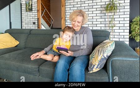 Großmutter und Enkelin verbringen Zeit miteinander und machen Online-Shopping auf einem Tablet. Die Großmutter sitzt mit dem Kind, während die Eltern bei Wor sind Stockfoto