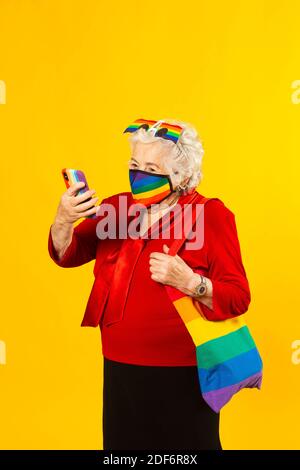 Studio-Porträt einer älteren Frau in einem roten Hemd, Regenbogen-Sonnenbrille, regenbogenfarbenen Gesichtsmaske und einer Tasche, mit einem Videoanruf mit ihrem Handy Stockfoto