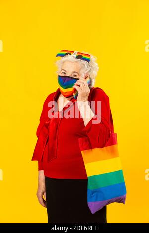 Studio Portrait einer älteren Frau in einem roten Hemd, Regenbogensonnenbrille, Regenbogenfarben Gesichtsmaske und einer Tasche, einen Anruf mit ihrem Handy, Stockfoto