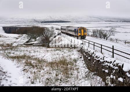Ribblehead, North Yorkshire. Dezember 2020. Die verschneite Szene in Ribblehead im Yorkshire Dales National Park. Ein Sprinter-Personenzug, der auf der berühmten Settle-Carlisle-Linie von Leeds nach Carlisle fährt, fährt über ein schneebedecktes Blea Moor, das gerade Ribblehead Viadukt überquert hat. Schwerer Schnee kam über Nacht auf dem höheren Boden in den Yorkshire Dales, einschließlich Ingleborough Peak, der in niedrigen Wolken im Hintergrund gesehen werden kann. Quelle: John Bentley/Alamy Live News Stockfoto