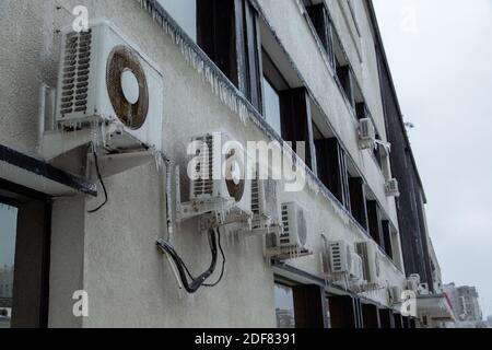 Viele weiße Klimaanlagen hängen in einer Reihe an einem grauen Gebäude mit verspiegelten Fenstern. Stark vereist. Stockfoto