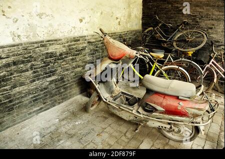 Alte kaputte und verlassenen Scooter durch Staub gegen verwitterte Wand geparkt. Rostiger Haufen von Fahrrädern im Hintergrund. Stockfoto