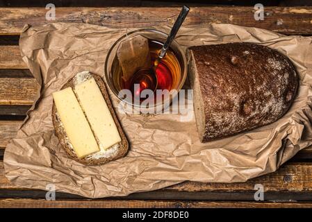 Brownbrot-Sandwich mit Käse und Butter, eine Tasse schwarzen Tee, ein halbes Laib braunes Brot auf Papiertüte. Stockfoto