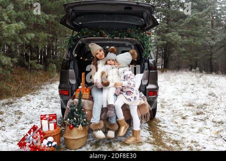 Eine junge Mutter und zwei kleine Mädchen sitzen im Kofferraum eines Autos und lachen fröhlich. Weihnachtsgeschenke im Wald in der Nähe des Autos. Frohe Weihnachten Stockfoto