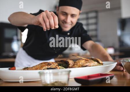 Männlicher kaukasischer Koch, der Salz über geröstete Fischgerichte zugibt Teller in der Restaurantküche Stockfoto