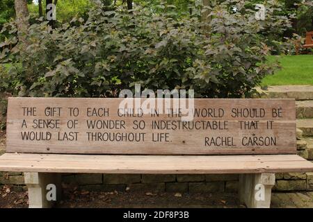 Eine Holzparkbank mit einem inspirierenden Zitat entlang einer steinernen Stützmauer, Sträuchern, Bäumen im Rotary Botanic Gardens in Janesville, Wisconsin, Stockfoto