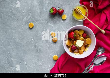 Trendiges Essen - Pfannkuchen Müsli. Haufen Mini-Cerealien-Pfannkuchen mit Erdbeeren und Honig. Draufsicht oder flach liegend, Kopierbereich. Stockfoto